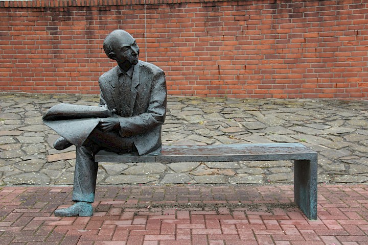 Statue: Mann sitzt auf Bank und hält eine Zeitung in der Hand